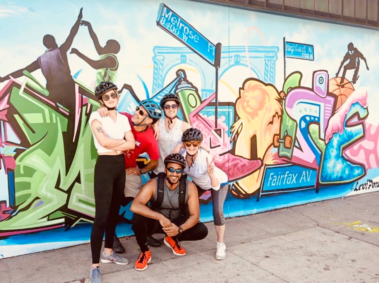 a photo of five people wearing sunglasses and bike helmets posing next to a mural of los angeles
