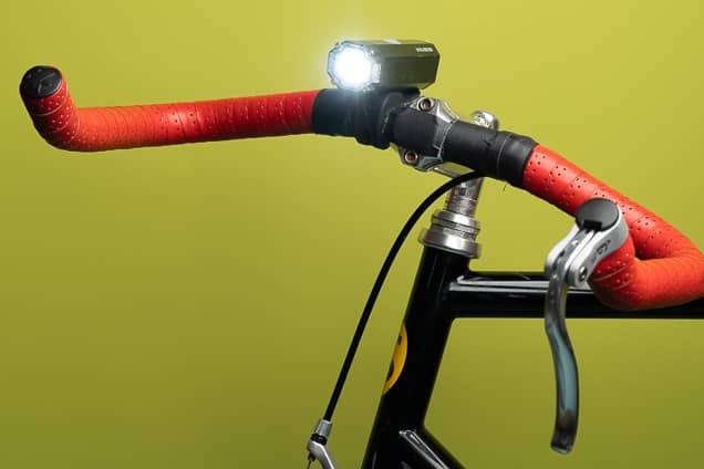 a close up of red bike handles on a black bicycle. there is an illuminated black bicycle light, and the photo has a chartreuse background.