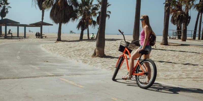 a woman riding an orange bike on a beach path near some palm trees